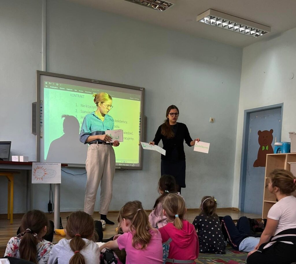 Prowadzące zajęcia pokazują kartki z emocjami. Dzieci siedzą na dywanie