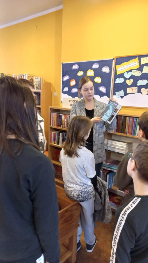 Na zdjęciu widoczna bibliotekarka i uczniowie. Bibliotekarka pokazuje uczniom książkę.