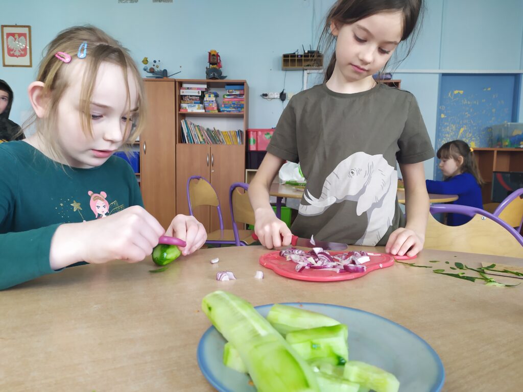 . Dziewczynki kroją cebulę oraz obierają zielonego ogórka