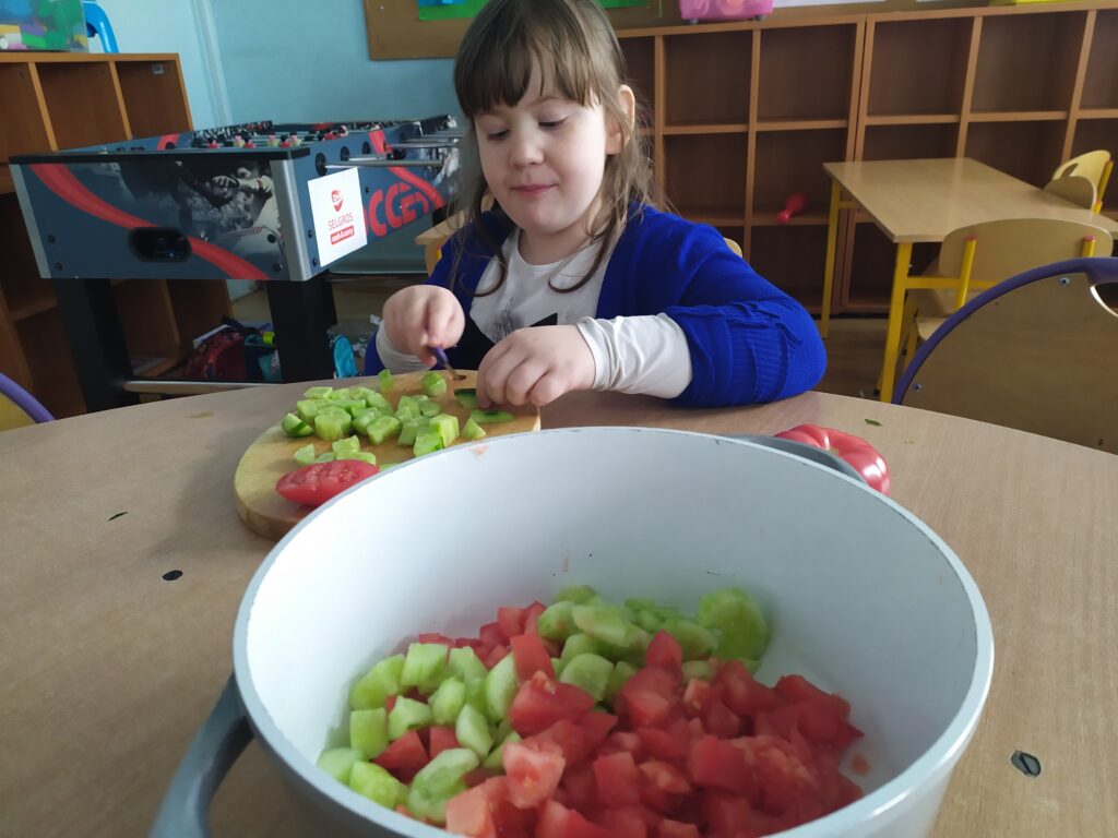 Miska z pokrojonymi warzywami, w tle uczennica krojąca zielonego ogórka.
