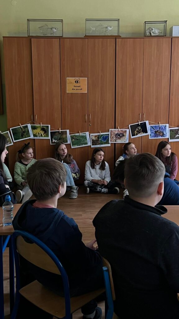 Uczniowie i uczennice siedzący na podłodze przy szafie. Nad nimi zdjęcia zapylaczy