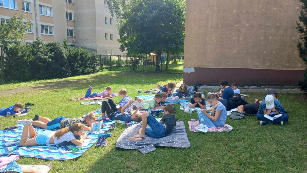 Uczniowie siedzą i leżą na kocach i czytają książki.