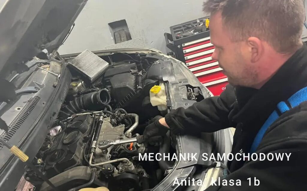 Zdjęcie przedstawia pracującego pod maską samochodową mechanika samochodowego.