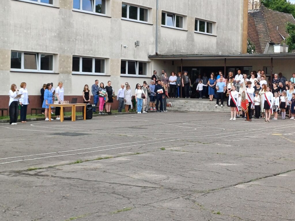 Nauczyciele wraz z uczniami i rodzicami na boisku szkolnym słuchają przemówienia.