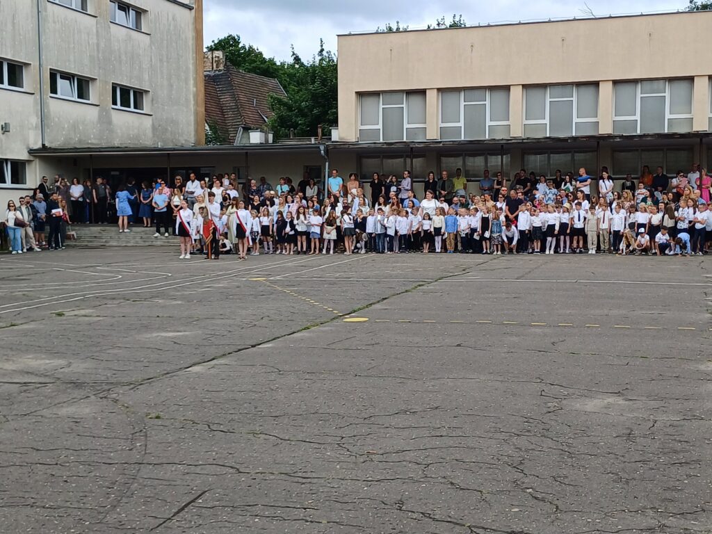 Zdjęcie przedstawia klasy pierwsze i drugie na boisku szkolnym w czasie apelu.
