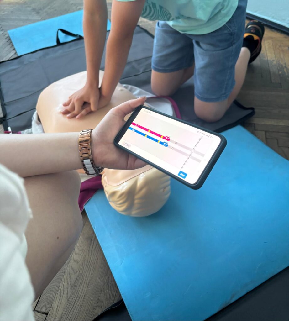 Telefon z aplikacją QCPR, a w tle dziecko cwiczy RKO