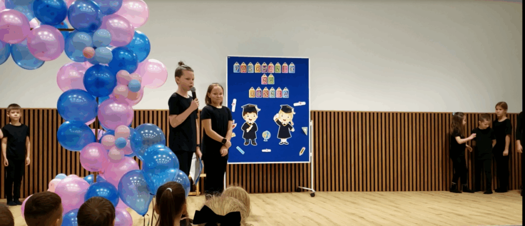 Uczeń i uczennica prowadzący uroczystość, stojący na scenie w auli szkolnej.