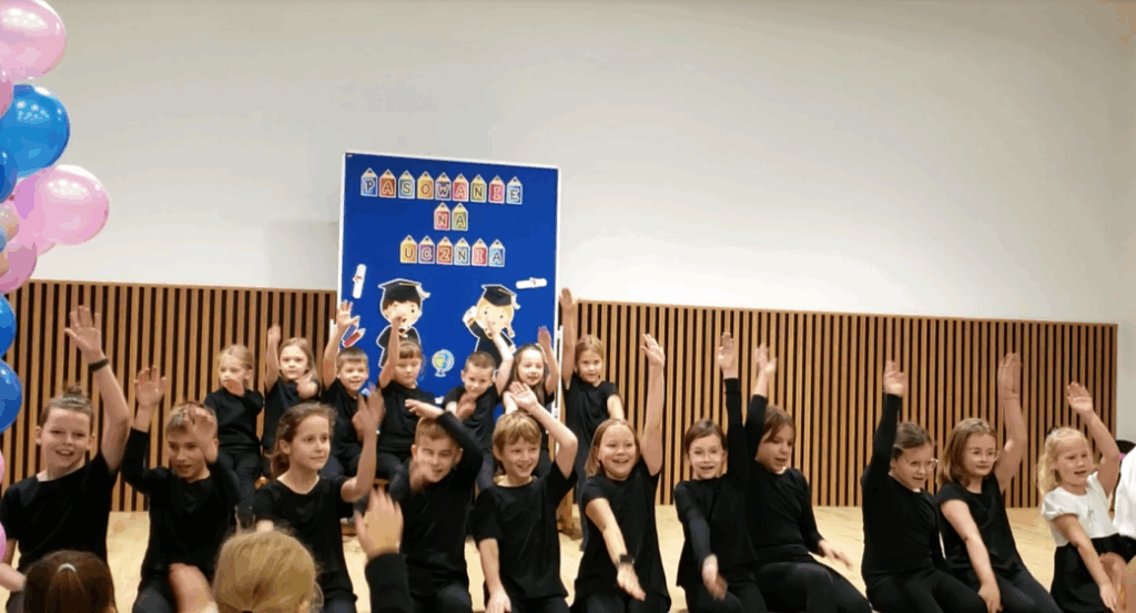 Wybrani uczniowie klas 1 oraz klasy 4c z koła artystycznego podczas tańca na scenie w auli szkolnej trzymając jedną rękę podniesioną do góry nad głowę.