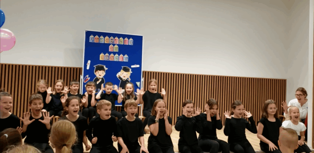 Niektórzy uczniowie z klas 1 i 4c z koła artystycznego tańczą na scenie - zdjęcie uchwyciło gestykulację/ imitację krzyku.