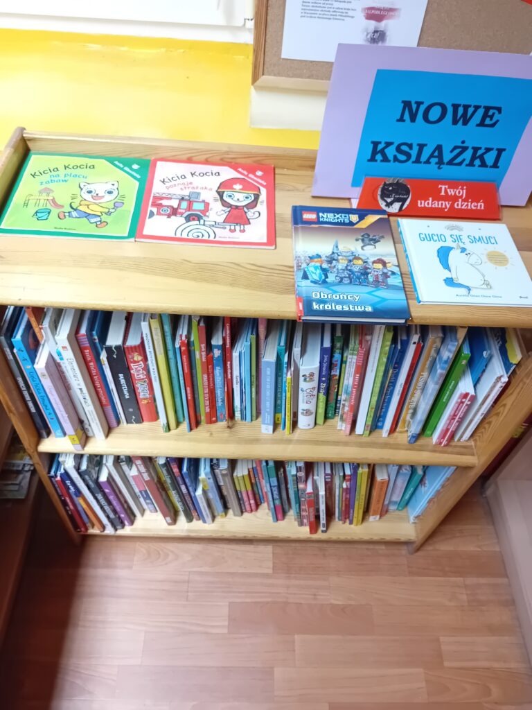 Książki stojące na półkach z informacją na górze na niebieskiej kartce, że są to nowe książki.