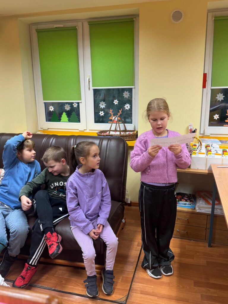 Uczniwoie siedzą na kanapie w bibliotece i słuchają uczennicy stojącej przy kanapie i czytającej tekst z kartki.