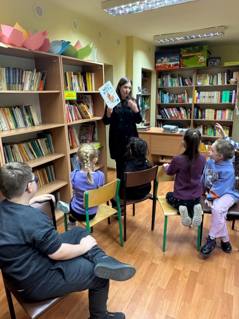 Uczniowie siedzący w bibliotece i słuchający Pani bibliotekarki.