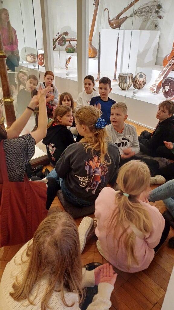 Uczniowie siedzący w pomieszczeniu na podłodze i oglądający, jak Pni prowadząca zajęcia prezentuje coś.