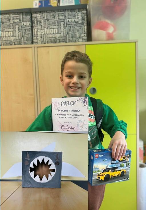 Uczeń z dyplomem i nagroda w dłoni stojący na rle szafek.