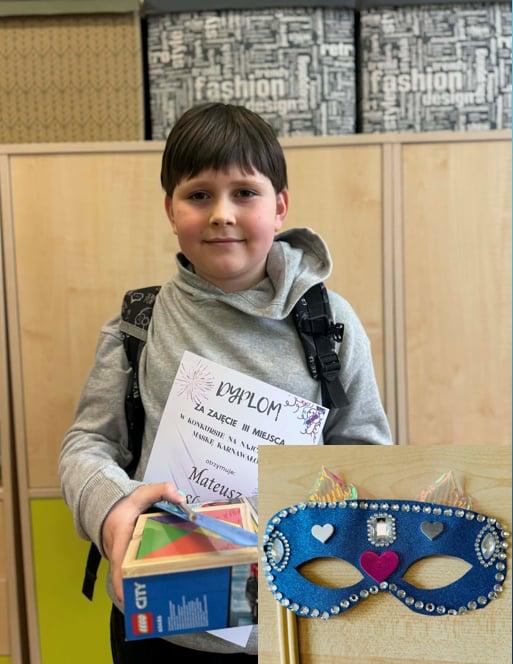 Uczeń stojący na tle szafek szkolnych w dłoni trzymający dyplom i nagrodę.