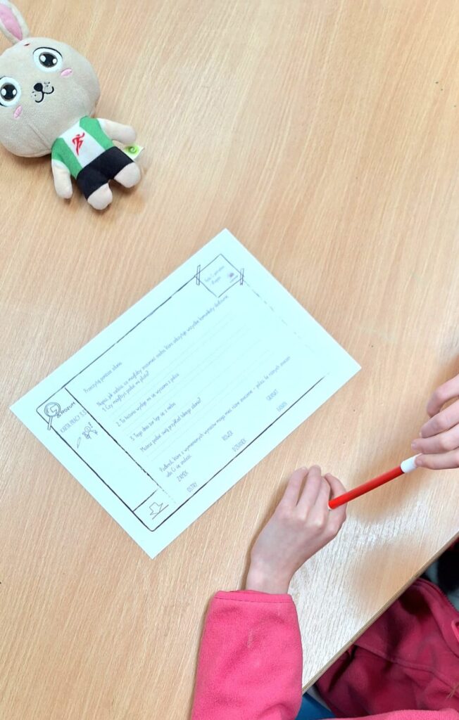 Na zdjeciu widać dłonie uczennicy na ławce szkolnej trzymające długopis oraz kartę pracy do wypełnienia.