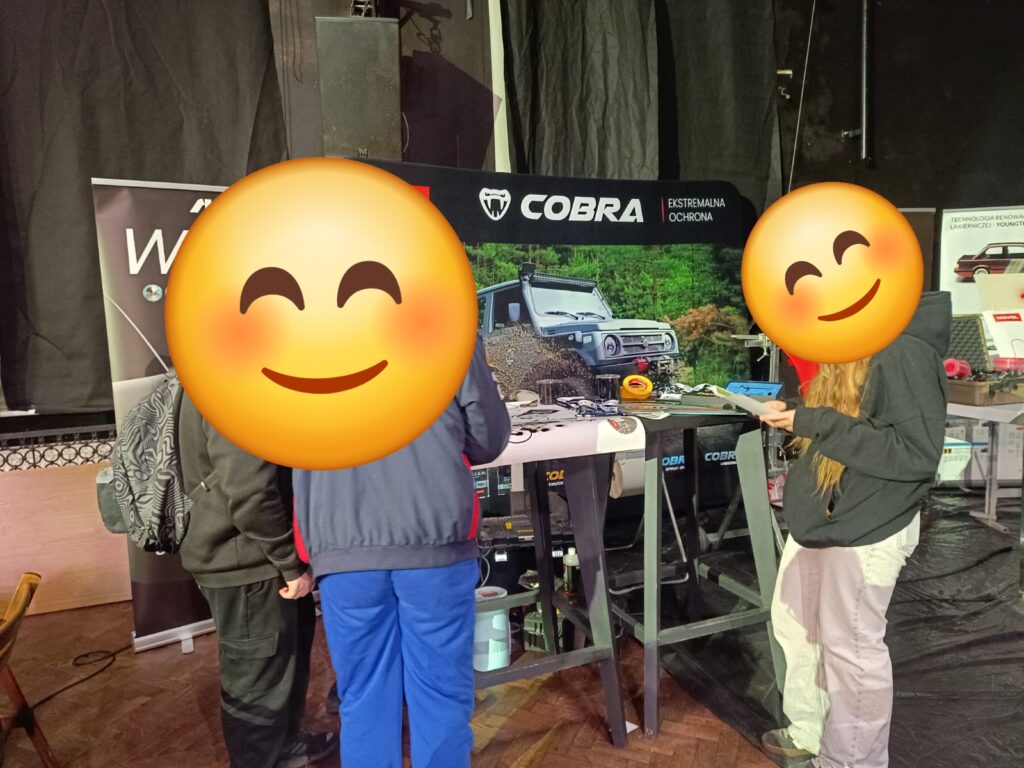 Zdjecie przedstawia osoby stojące przy stoiku i oglądające wystawę.