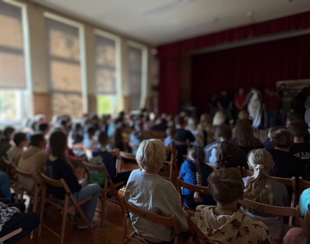 Zdjęcie przedstawia uczniów siedzących w auli szkolnej podczas występów.